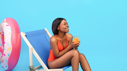 Cheerful black woman relaxing in lounge chair with fruit cocktail, laughing and enjoying her...