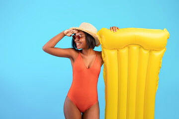 Young black lady in swimsuit with inflatable lilo holding hand above her eyes, looking into distance on blue background