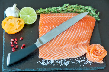 Hand chef using knife slice raw salmon on chopping block. Salmon fillet steak and seasonings on black cutting board. 