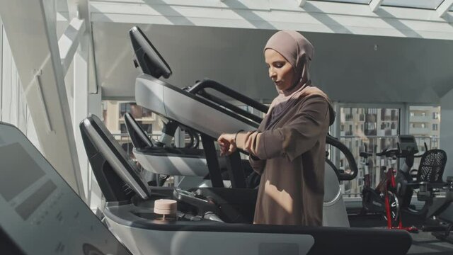Young Muslim woman in hijab running on treadmill, then pressing stop button and drinking water after gym workout