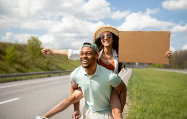 Obraz premium Happy young multiracial couple hitchhiking together, holding empty cardboard sign, showing thumb up gesture outdoors