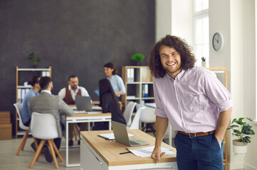 Cheerful businessman on foreground looking at camera with smile standing in office with colleagues sitting and talking brainstorming at table. Glad to be on team, coworking space concept