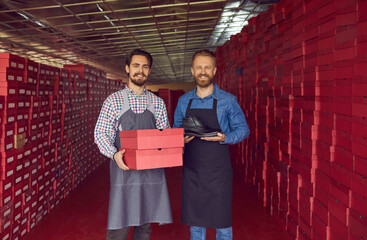 Two happy proud shoe factory workers holding red cardboard boxes and showing new male leather boot...