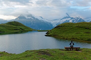 Tourist sitting on a bench by the grassy lakeside of Bachalpsee & enjoying the panoramic view of majestic alpine mountains under moody cloudy sky in First, Grindelwald, Bernese Highlands, Switzerland