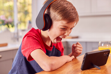 Boy At Home In Kitchen Watching Movie Or Show On Mobile Phone Wearing Wireless Headphones