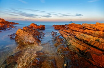 Sunrise scenery of a beautiful rocky beach in northern Taiwan, with golden sunlight illuminating the rocks and an island on distant horizon under dramatic dawning sky (Long Exposure Effect )