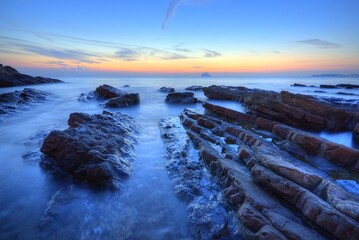 Beautiful sunrise scenery of a rocky beach on northern coast of Taiwan with an island on distant horizon & peculiar rock formations on seashore under dramatic dawning sky (Long Exposure Effect)