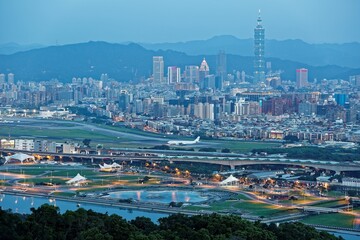 Aerial panorama of Taipei in evening twilight, capital city of Taiwan, with Taipei 101 Tower amid skyscrapers in downtown, a riverside park by Keelung River & an airplane parking in Songshan Airport