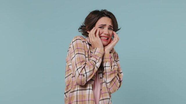 Scared shocked sad young brunette woman 20s years old wears plaid shirt look camera covering hiding face with hands peep through fingers isolated on pastel plain light blue background studio portrait