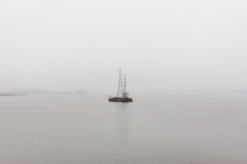 Power lines on small island on river. Electricity tower with wires in mist. Foggy autumn day.