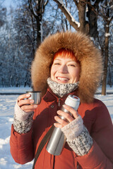 Smiling woman in red coat drinks hot tea from metal thermos. Leisure activity in winter forest at sunset.