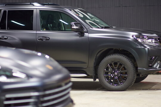 Toyota Land Cruiser And Porsche Cayenne Cars Covered With A Protective Matte Paint Film In Black Are In The Repair Shop, Blurred Focus. Chelyabinsk, Russia, April 03, 2021