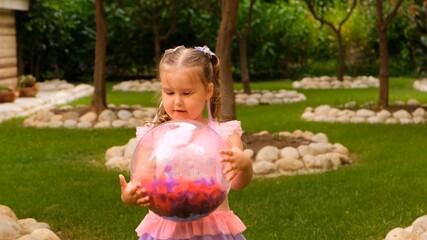 cute little girl, 3 years old, with two ponytails on her head, dressed in a delicate and multi-colored dress of pink blue color, plays with a bright transparent ball with multi-colored feathers inside