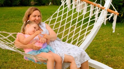 Grandmother and granddaughter, 3 years old, rest and bask in a hammock on a green meadow in their garden on a sunny summer day. Vacation concept, generational relationship, parenting, happy childhood.