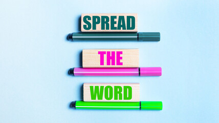 On a light blue background, there are three multi-colored felt-tip pens and wooden blocks with the SPREAD THE WORD