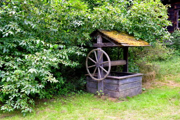A close up on an old, abandoned and damaged well with some moss on its roof and with a small wooden wheel located next to a dense forest or moor seen on a Polish countryside in summer