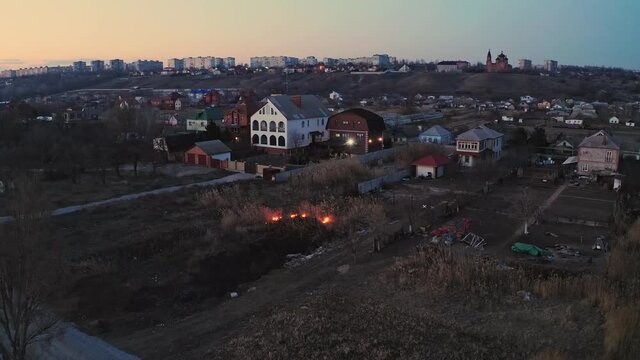 Evening fire in a field near the city. Burning reeds near houses. Seasonal fires in the steppe area. Fire element as a consequence of arbitrary bonfires. Drone video.
