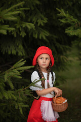 A cute little girl in the image of little red riding hood is standing in a spruce forest