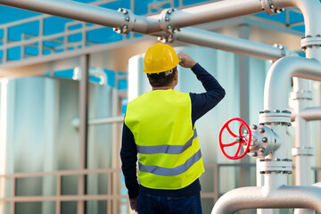 A worker in a helmet and a yellow vest next to a valve against the background of an oil and gas...
