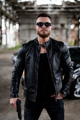 Man with a beard in a black leather jacket, wearing sunglasses and holding a gun. Man is looking at the camera. Guns concept