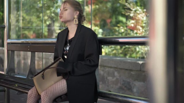 Anxious stressed worried woman with empty purse sitting on bus stop outdoors. Portrait of young beautiful Caucasian party-goer robbed in urban city in the morning. Crime and lifestyle