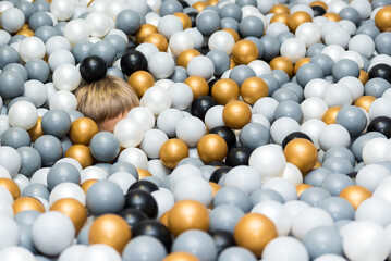 Many different color (white, gold, grey, black) plastic balls and kid's hair is visible in ball pool at indoors playground