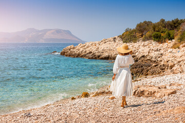 Happy and elegant woman traveler in hat and a charming white dress is walking on the sea shore of a...