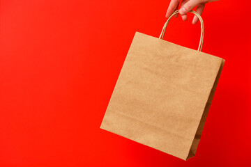 Female hand holding brown craft blank shopping bag isolated on red background. Black friday sale, discount, recycling, shopping and ecology concept.
