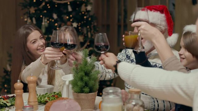 Family New Years Dinner. At Table An Elderly Man In Santa Hat And Woman With Glasses. Two Children Boy And Girl And Two Young People. They Knock Over Glasses And Celebrate.