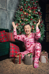 Cheerful surprised funny man in pink sleepwear with hands up sitting near decorated fir tree and...