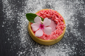 Cake with cream and white flower on a black surface, sprinkled with powdered sugar.