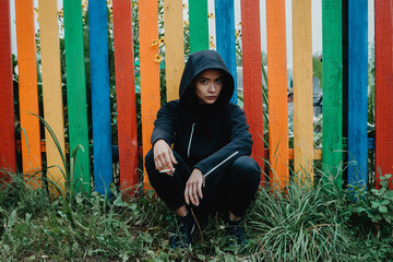 Beautiful young woman in a hood sits on his haunches with a cigarette in her hand near a...