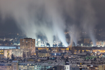 Night city view with buildings and a smoking industrial steel mill with impressive exhaust gases