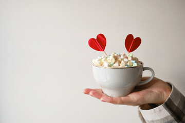 A cup of hot chocolate with tender marshmallows and two hearts in hand on a light background. The concept of sweet love, Valentine's day, romance. Festive sweets close-up