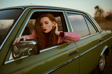 Fashionable redhead woman looking in the mirror, posing in the green vintage car. Copy, empty space for text

