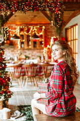 beautiful young woman in red pajamas sits near Christmas tree in living room decorated for celebration of Christmas and New Year and opens gift box with wrapped Christmas presents