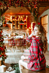 beautiful young woman in red pajamas sits near Christmas tree in living room decorated for celebration of Christmas and New Year and opens gift box with wrapped Christmas presents