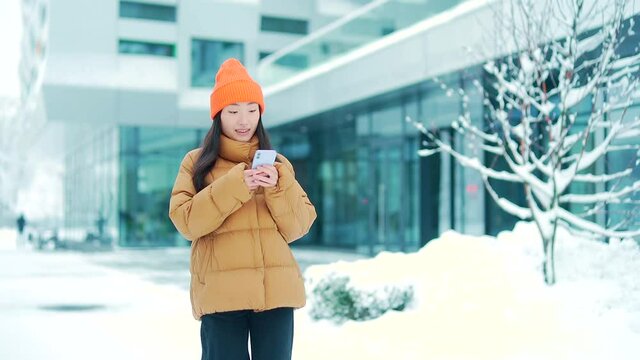 Beautiful happy asian teen girl or student walks on winter snowy city streets in cold weather uses mobile phone. Smile. Young woman with smartphone in hands scrolling texting using. Outside, outdoors