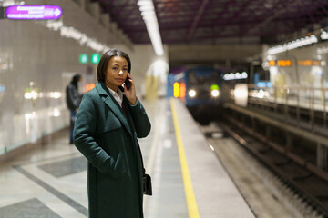 Young african american woman office worker making phonecall while standing at subway platform after workday and waiting for train. African female in coat talking by mobile phone at underground station