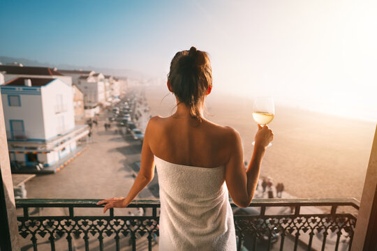 Woman wrapped in towel standing on the balcony and holding glass of white wine in luxury apartment with ocean view. Woman on vacation relaxing and enjoying life