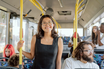 Smiling, attractive student in strange city, moving by public transport, girl is holding on to the railing, looking into the camera with joy, around other passengers traveling by bus to work, school