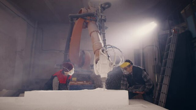 Zoom out view of man and woman in respirators and face mask discussing robotic arm milling machine cutting block of polymer in workroom