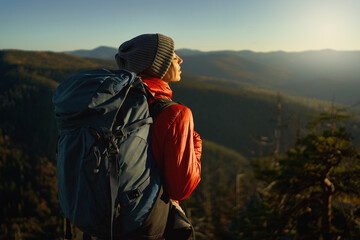 Back view woman hiker in bright red jacket with backpack standing on edge of cliff against background of sunset sky over mountains