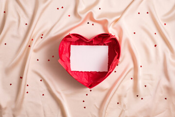 Open heart shaped box with blank paper card mockup inside on beige textile background.