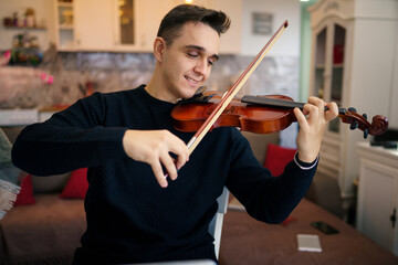 One young adult caucasian man sitting on the chair at home learning to play violin male violinist...