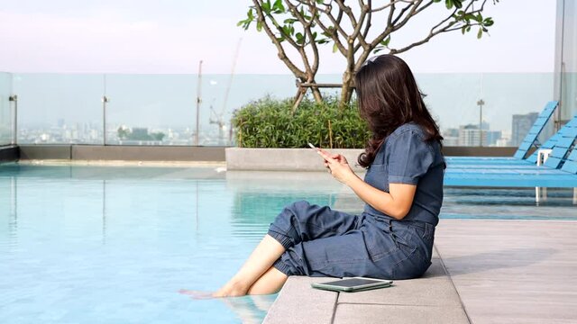 Asian woman using smartphone share photos, communicate with friends on vacation, sitting on the edge dangling her feet in swimming pool on skyscraper, woman looking at cityscape while relaxing. 