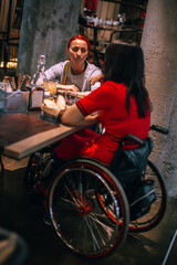 Obraz premium Two girls in wheelchairs in cozy cafe waiting for order. Accessible common areas for people with special needs. Affordable tourism for people with special needs due to physical special needs