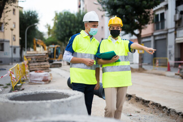 Male and female european builders in face masks talking about their work while standing in construction plant. Woman pointing with finger.