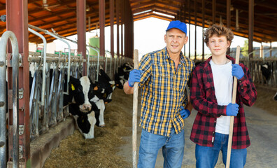 Adult and young male farmers with working tools in hands standing in cowshed and looking in camera.