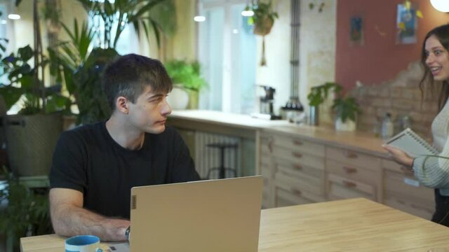 Young european male Student is at the coffeehouse with natural background. Black hair female asking to work together. Sitting at the table and showing on the laptop smiling and talking. Slow Motion.